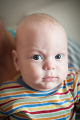 A young baby with blue eyes and a slightly skeptical expression looks directly at the camera. The baby is wearing a colorful striped shirt.