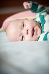 A young baby is lying on its back on a light blue blanket. The baby is wearing green and white striped pajamas and has its mouth open while looking up.