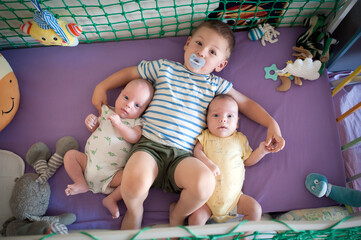 An older brother cuddles with his newborn twin brother and sister on a purple crib mattress surrounded by colorful plush toys. The scene occurs indoors during the daytime.