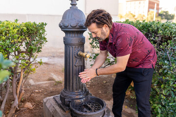 Man with dreadlocks washing hands under fresh water from an outdoor iron fountain