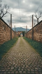 Obraz premium Cobblestone path leads to an old archway through ancient stone walls, flanked by bare trees and utility poles, under a dramatic cloudy sky with distant mountains.