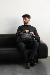 A professional male model in charcoal knitwear and tailored trousers sits on a black leather sofa holding a premium black leather briefcase.