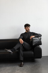 A professional male model in charcoal knitwear and tailored trousers sits on a black leather sofa holding a premium black leather briefcase.