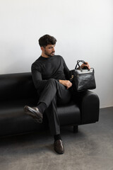 A professional male model in charcoal knitwear and tailored trousers sits on a black leather sofa holding a premium black leather briefcase.
