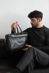 A professional male model in charcoal knitwear and tailored trousers sits on a black leather sofa holding a premium black leather briefcase.