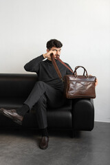 A professional male model in charcoal knitwear and tailored trousers sits on a black leather sofa holding a premium brown leather briefcase.