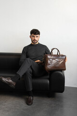 A professional male model in charcoal knitwear and tailored trousers sits on a black leather sofa holding a premium brown leather briefcase.