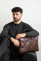 A professional male model in charcoal knitwear and tailored trousers sits on a black leather sofa holding a premium brown leather briefcase.