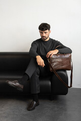 A professional male model in charcoal knitwear and tailored trousers sits on a black leather sofa holding a premium brown leather briefcase.