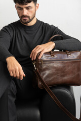 A professional male model in charcoal knitwear and tailored trousers sits on a black leather sofa holding a premium brown leather briefcase.