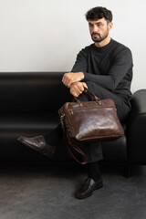A professional male model in charcoal knitwear and tailored trousers sits on a black leather sofa holding a premium brown leather briefcase.