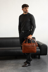 A professional man in a charcoal crewneck sweater and tailored trousers holds a light brown briefcase standing in a minimalist studio.