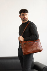 Professional male model in charcoal crewneck knitwear holding a light brown leather briefcase in a minimalist studio setting