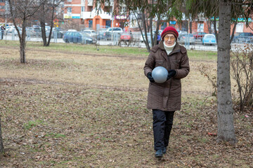 Elderly woman with a red hat holding a ball, walking in an autumn park. Healthy active life, retirement, and social care concept for nursing home.