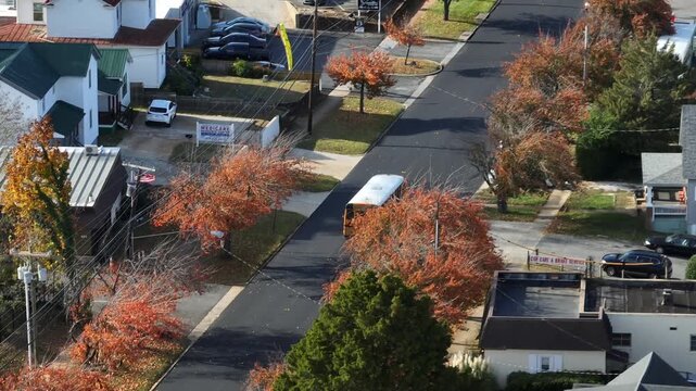 Aerial view of yellow school bus driving through an American residential neighborhood in autumn, with colorful fall trees, quiet streets and suburban houses under clear daylight. tracking shot.