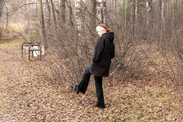Young european woman with red hair walking away in an autumn forest with dry leaves on the ground and bare trees, thoughtful.