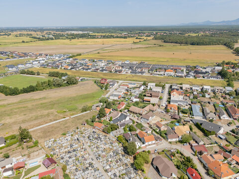 Aerial view of a cemetery with rows of headstones contrasting with the adjacent residential area and golden fields under a clear sky, Lab, Bratislava Region, Slovakia.
