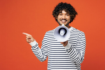 Young Indian man wear striped sweatshirt casual clothes hold megaphone scream announces discounts sale Hurry up point aside isolated on plain red orange background studio portrait. Lifestyle concept.