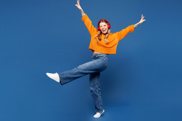 Full body young happy smiling Caucasian woman she wear yellow sweater casual clothes listen to music in headphones raise up hands isolated on plain blue background studio portrait. Lifestyle concept.