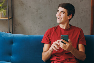 Fototapeta premium Young cheery man wear red t-shirt look aside hold use mobile cell phone sit on blue sofa stay at home hotel flat rest relax spend free spare time in living room indoor grey wall People lounge concept