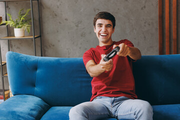 Young excited man wear red t-shirt hold play pc game with joystick console sit on blue sofa couch stay at home hotel flat rest relax spend free spare time in living room indoors People lounge concept. © ViDi Studio