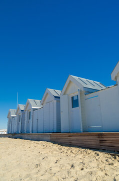 View of a row of white beach huts standing proudly against the vast blue sky, their simple architecture a stark contrast to the soft, inviting sand, Arcachon, France.