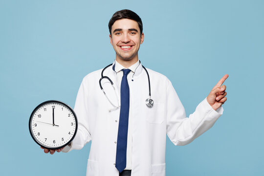 Young male doctor man wear white medical gown suit work in hospital clinic office hold in hands clock show time point finger aside isolated on plain blue background studio Healthcare medicine concept