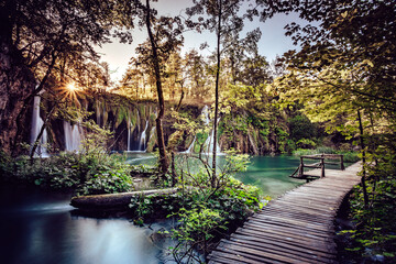 View of wooden walkway leading to turquoise waters beneath cascading waterfalls, sunlight piercing through lush trees, Plitvicka Jezera, Lika-Senj County, Croatia.