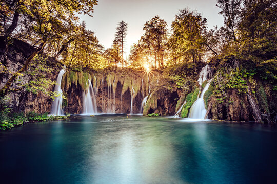 View of sunlight streams through the trees, illuminating the cascading waterfalls and the serene turquoise lake below, Plitvicka Jezera, Lika-Senj County, Croatia.