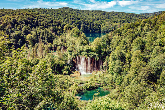 View of cascading waterfalls plunge into the turquoise pools surrounded by lush green forests under a bright blue sky, Plitvicka Jezera, Lika-Senj County, Croatia.