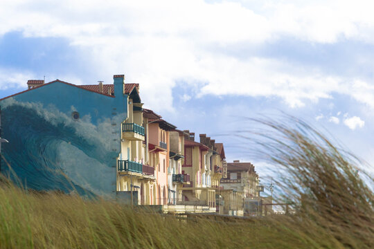 View of pastel buildings adorned with a striking wave mural stand against a cloudy sky as windswept grasses dance in the foreground, Hossegor, Nouvelle-Aquitaine, France.