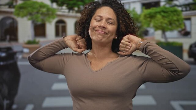 Hispanic middle aged woman stretching neck and smiling under afternoon sunlight on a busy street; serenity.
