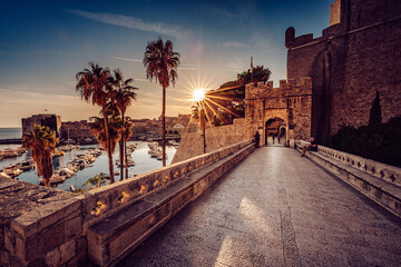 View of the stone bridge leading to the ancient city gates, bathed in the warm glow of the setting sun, palm trees sway gently in the breeze, Dubrovnik, Dubrovnik-Neretva County, Croatia.