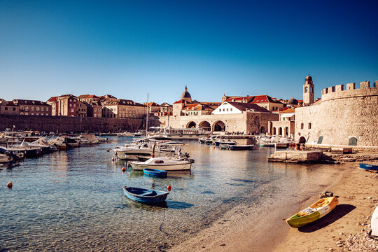View of boats bob gently in the harbor, reflecting the clear sky as stone walls and buildings glow under the warm sunlight, Dubrovnik, Dubrovnik-Neretva County, Croatia.