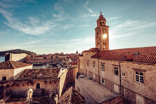 View of aged stone buildings with red tile roofs bask under a sun-drenched sky, the tower clock face reflecting light in old town, Dubrovnik, Dubrovnik-Neretva County, Croatia.