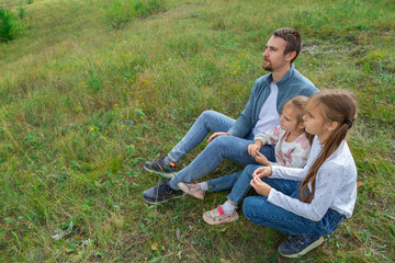 Fototapeta premium Father and daughters sitting on top of hill and enjoy beautiful scenery of green hills under blue cloudy sky.