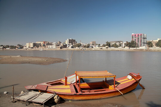View of a vibrant orange and red boat resting on the muddy bank, mirroring the cityscape across the serene water in Sukkur, Sindh, Pakistan.