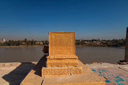 View of a carved stone structure with Arabic script stands prominently against the backdrop of a wide, calm river, under a clear blue sky, Sukkur, Sindh, Pakistan.