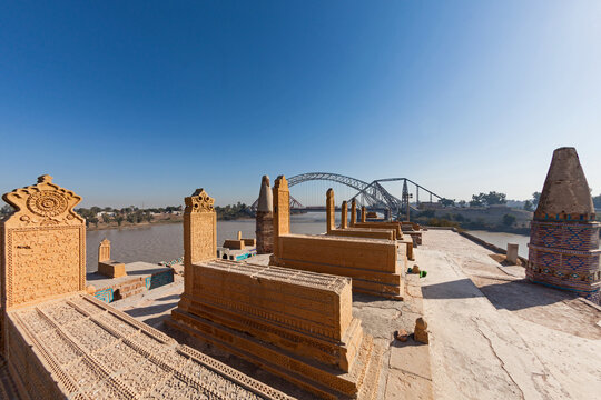 View of intricately carved tombs casting long shadows under a clear sky, with a modern arch bridge spanning the wide river, Sukkur, Sindh, Pakistan.