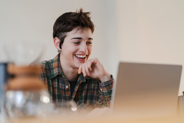 A young person with short hair smiles and laughs, looking at a laptop screen. They are enjoying engaging digital content or a virtual connection in a bright indoor setting.