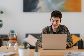 A young person with short hair and earbuds laughs while working on a laptop at a wooden table. They wear a plaid shirt. Coffee tools and plants are on the desk.