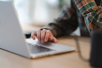 A person in a plaid shirt uses a laptop on a wooden desk. Their hand is on the trackpad, navigating or working. This image conveys focus, remote work, or casual computing.