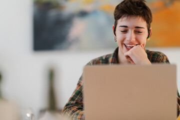A smiling young person with short hair is focused on their laptop screen. They wear a plaid shirt and earbuds, enjoying an online activity indoors.