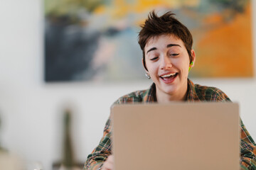 A happy young person with short hair and an earbud smiles broadly, looking at their laptop screen. They are wearing a plaid shirt, enjoying a moment of online engagement.