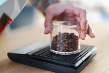 A person's hand carefully places a clear container filled with dark roasted coffee beans onto a digital kitchen scale. This precise weighing is essential for quality coffee preparation.
