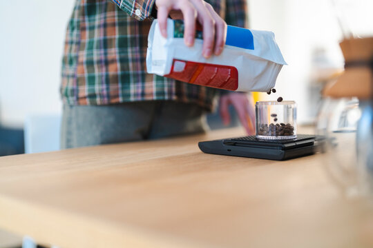 A person in a plaid shirt pours whole coffee beans from a bag into a clear container resting on a digital scale. This action demonstrates precise coffee preparation.