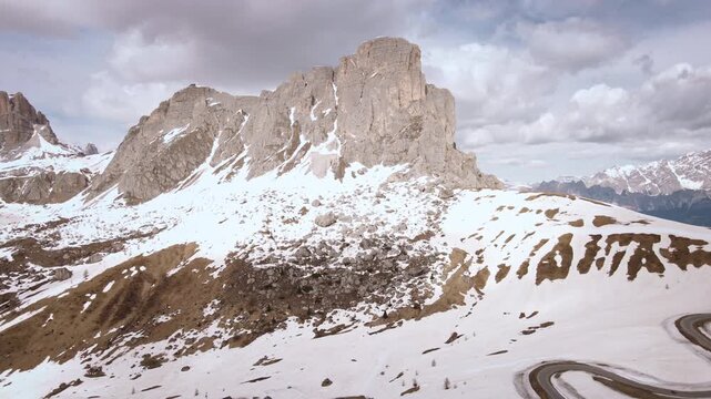 Aerial camera moves backward from Ra Gusela peak, revealing snowy slopes and winding Passo Giau road in the Dolomites, Italy, under dramatic clouds.