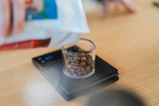 A person's hand carefully pours roasted coffee beans from a white bag into a clear glass container resting on a digital scale. This precise measurement is essential for brewing quality coffee.