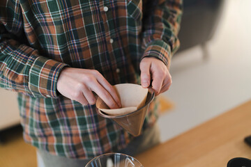 A person in a green and orange plaid shirt prepares to brew fresh coffee. They are carefully inserting a brown paper filter into a pour-over coffee dripper, ready for the brewing process.