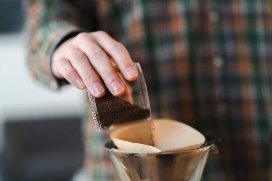 A person's hand pours ground coffee from a clear container into a paper filter inside a metal pour-over coffee maker. This is part of the coffee brewing process.